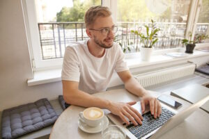 portrtait-young-cute-guy-working-remotely-city-cafe-looking-away-with-gentle-smile-making-pause-with-work-thinking-about-something-positive - SIM Computer W dzisiejszych czasach laptop to nieodzowny element wyposażenia każdego profesjonalisty.